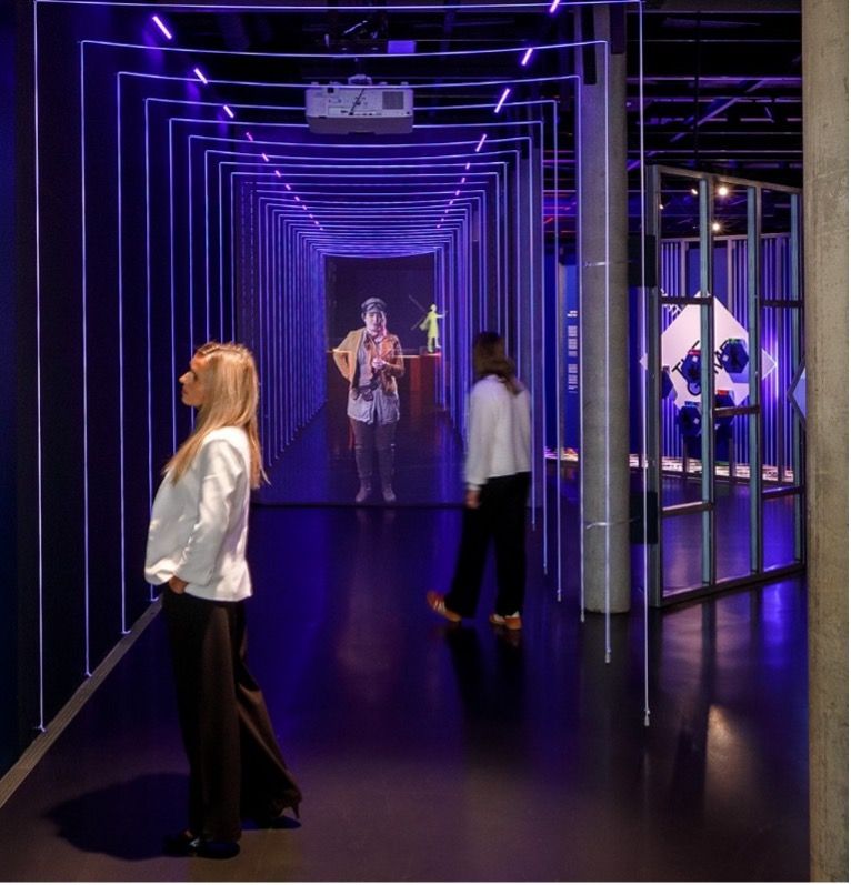 A modern museum exhibit features a tunnel of glowing purple LED frames, leading to a holographic-style projection of a historical figure. Two visitors stand observing the installation. On the right, another part of the exhibit displays objects in a glass case under similar purple lighting. The setting combines immersive technology with historical storytelling.