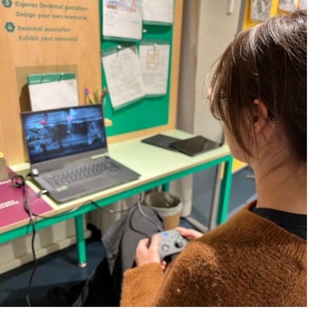 A person with brown hair, wearing a brown sweater, is playing a historical video game on a laptop using a game controller. The screen shows a black-and-white scene, likely from Through the Darkest of Times. The setup is on a green table in an educational or museum setting, with informational materials pinned on a green bulletin board in the background.